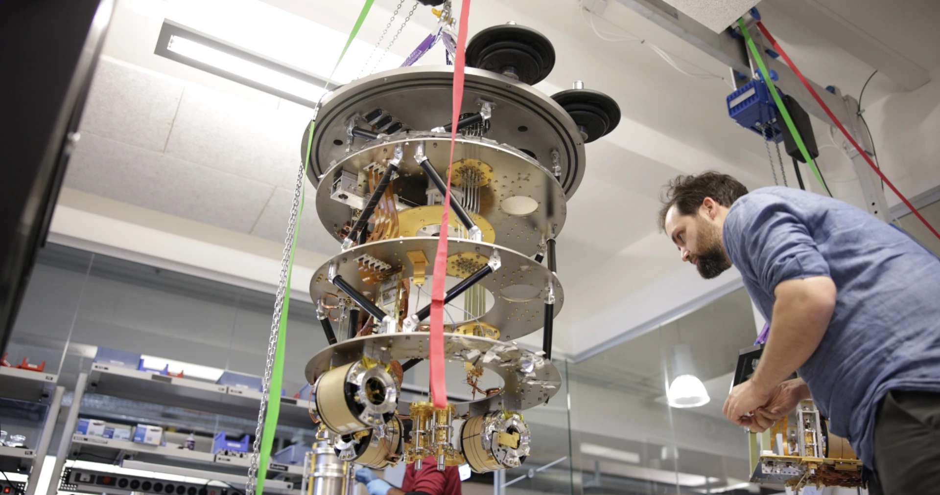 A kiutra cryostat photographed from below, its color-coded wiring loom visible, with an engineer working beside it in the lab.