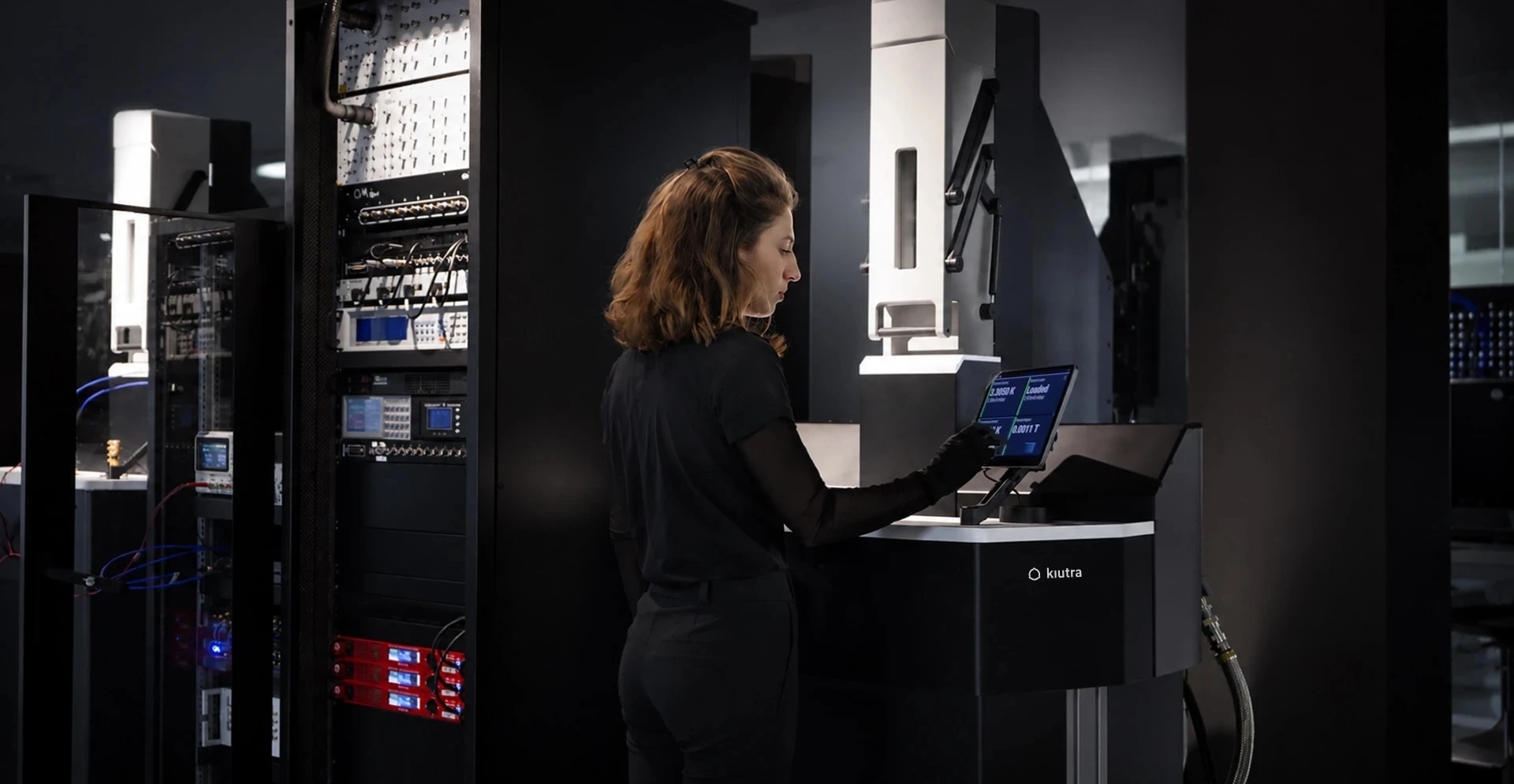 A kiutra researcher operating an L-Type Rapid cryostat from the tablet mounted to the system, with measurement racks and a second system in the kiutra lab