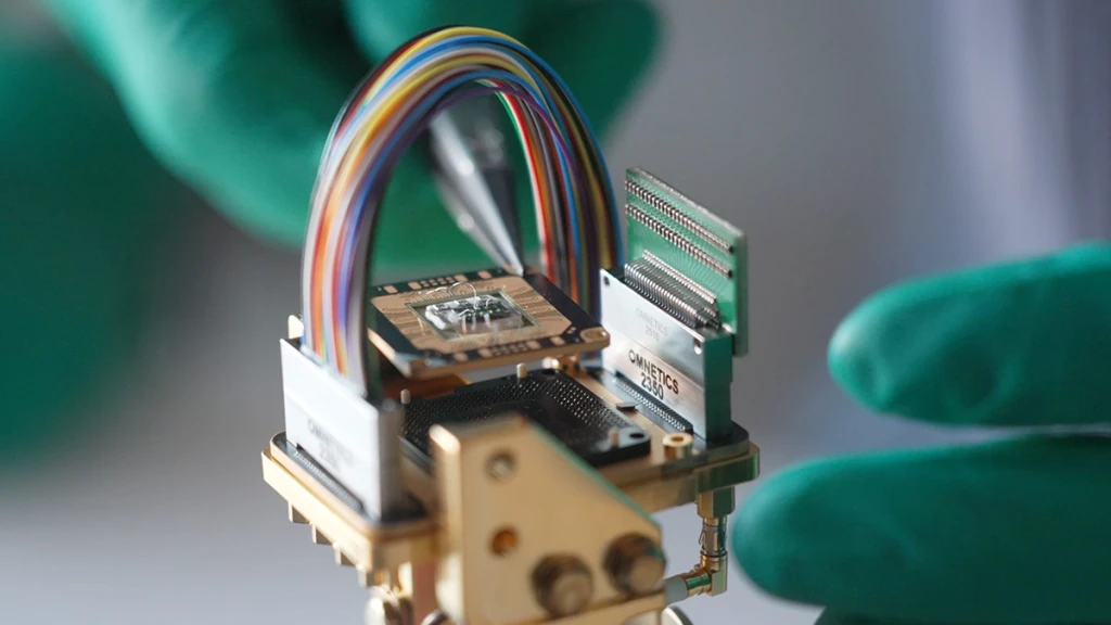 Close-up of a gold-plated sample holder held in a gloved hand: a wire-bonded chip on a carrier board between two Omnetics connectors, a ribbon cable arching over it.