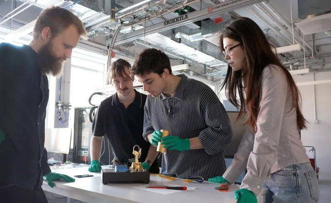 Four people in a university lab around a bench, one of them in green gloves holding a gold-plated sample puck above the puck station.