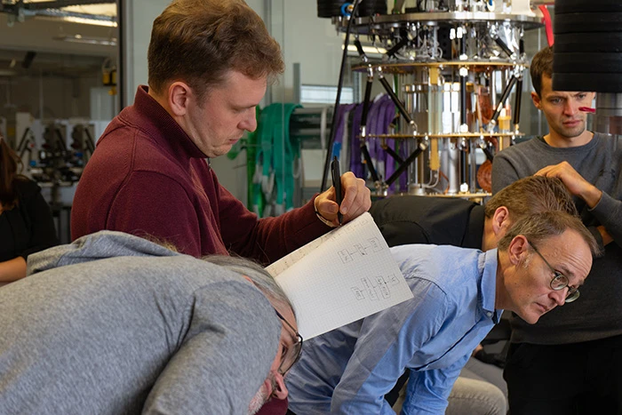 Four researchers leaning over a notepad sketch beside a cryostat in the kiutra lab during the scesamm workshop
